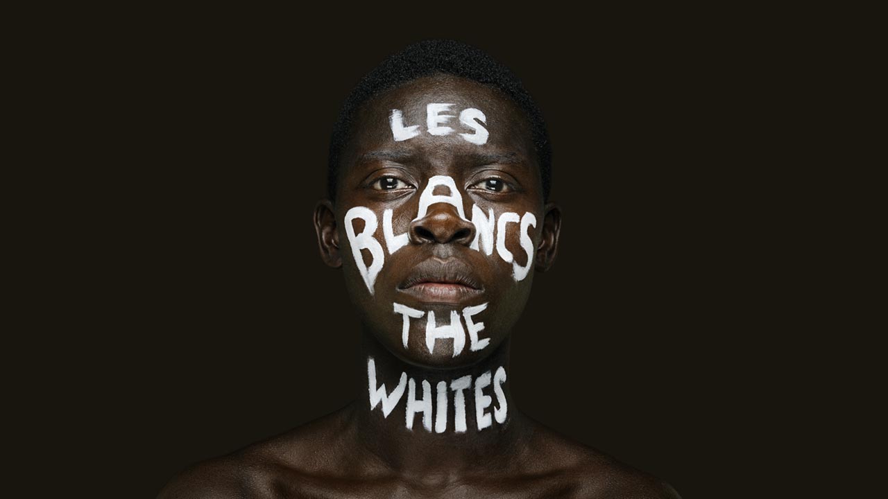 A photo of a Black man's face with the words Les Blancs | The Whites witten on his face in white paint. A Black man;s face with the words— Les Blancs The Whites— witten on his face in white paint.